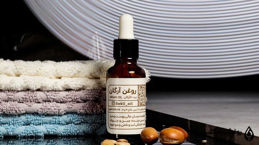 یک شیشه حاوی روغن آرگان در کنار یک حوله و چند دانه آرگان