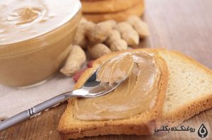 یک قاشق بر روی نان تست حاوی کره بادام زمینی دانه های بادام زمینی و یک ظرف حاوی کره بادام زمینی